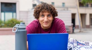 A student smiling on campus with a laptop and water bottle.
