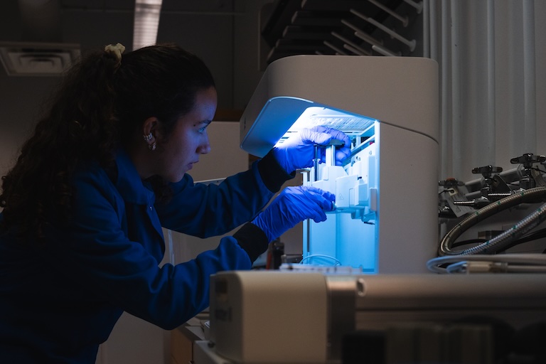 A person in a blue lab coat and purple gloves operates laboratory equipment illuminated with blue light. The individual’s face is obscured. Additional lab equipment and cables are visible in the background.