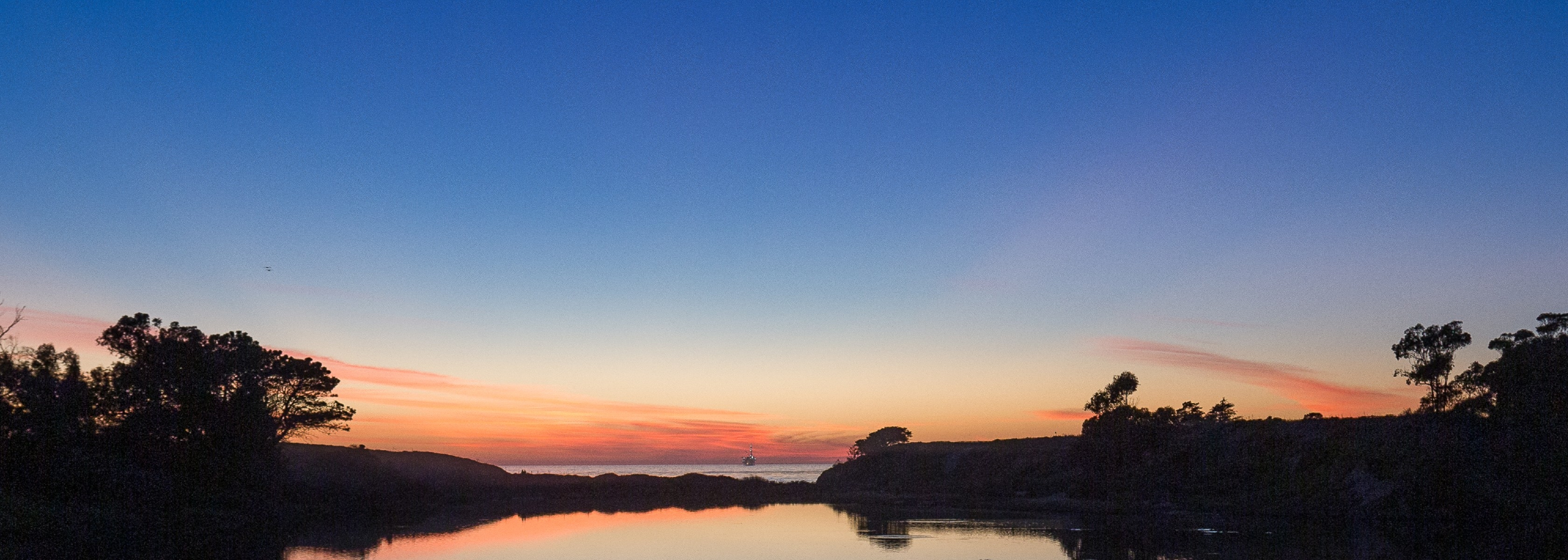 A wide panoramic view of the UC Santa Barbara lagoon at twilight, with deep blue water reflecting the oranges and pinks of a setting sun.