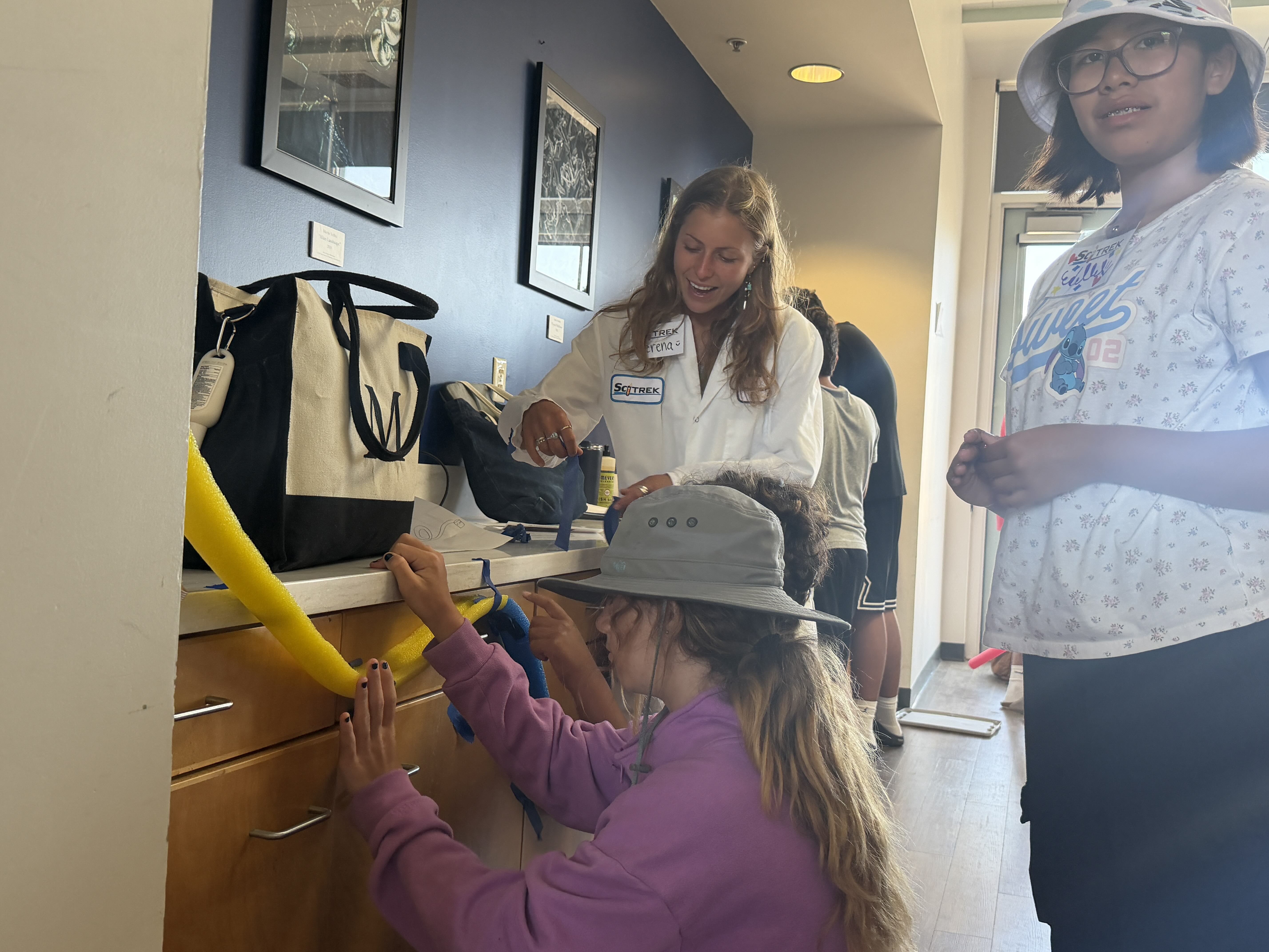 A SciTrek mentor talks with campers as they adjust a yellow pool-noodle roller coaster attached to a cabinet.