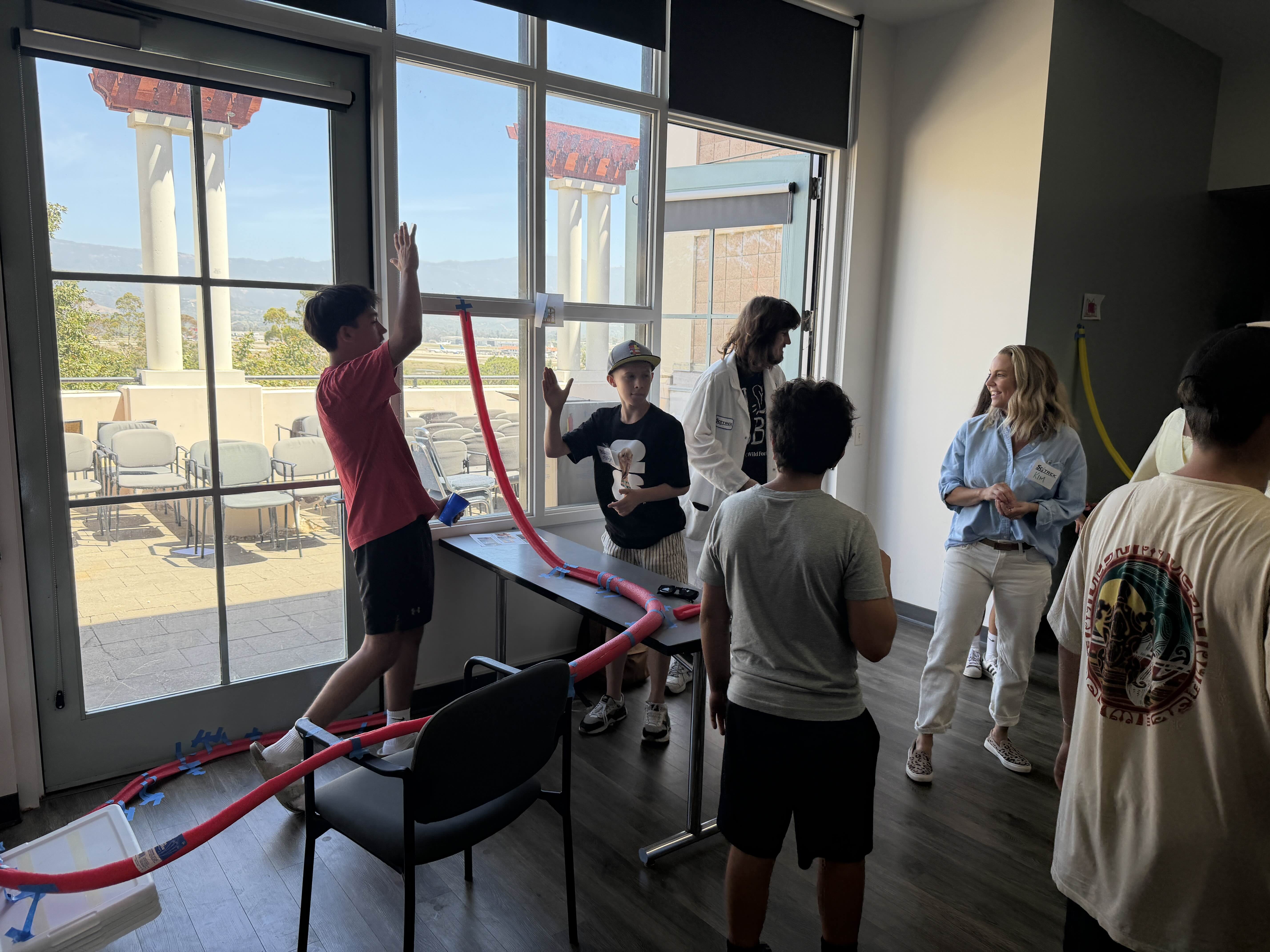 A camper raises a hand beside a red pool-noodle roller coaster while SciTrek mentors and other campers gather nearby.