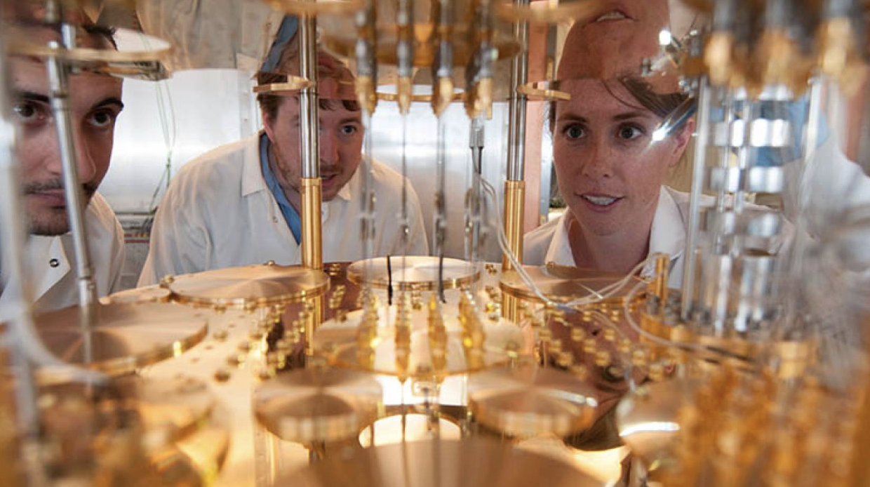 Three scientists, two men and one woman, all wearing white lab coats, lean in close to observe and work on a complex, highly polished, gold-colored scientific apparatus with multiple reflective layers and components in a laboratory setting. Their faces are partially visible and focused on the center of the machine.
