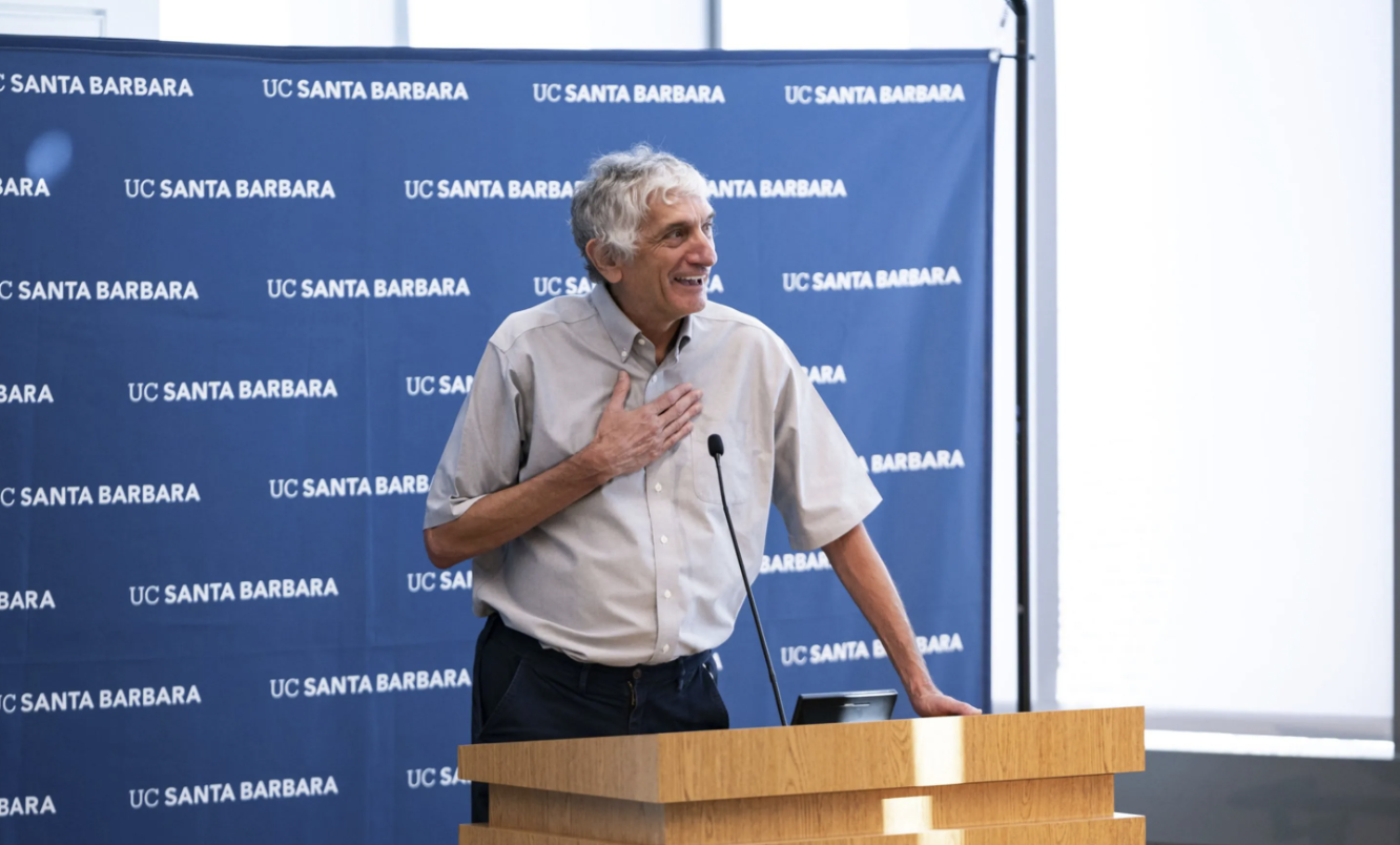 A smiling, older man with gray hair is speaking at a light wood podium during a presentation. He is wearing a light gray short-sleeved collared shirt and dark pants, and has one hand resting on his chest. Behind him is a dark blue backdrop covered with a repeating pattern of the white text UC SANTA BARBARA.