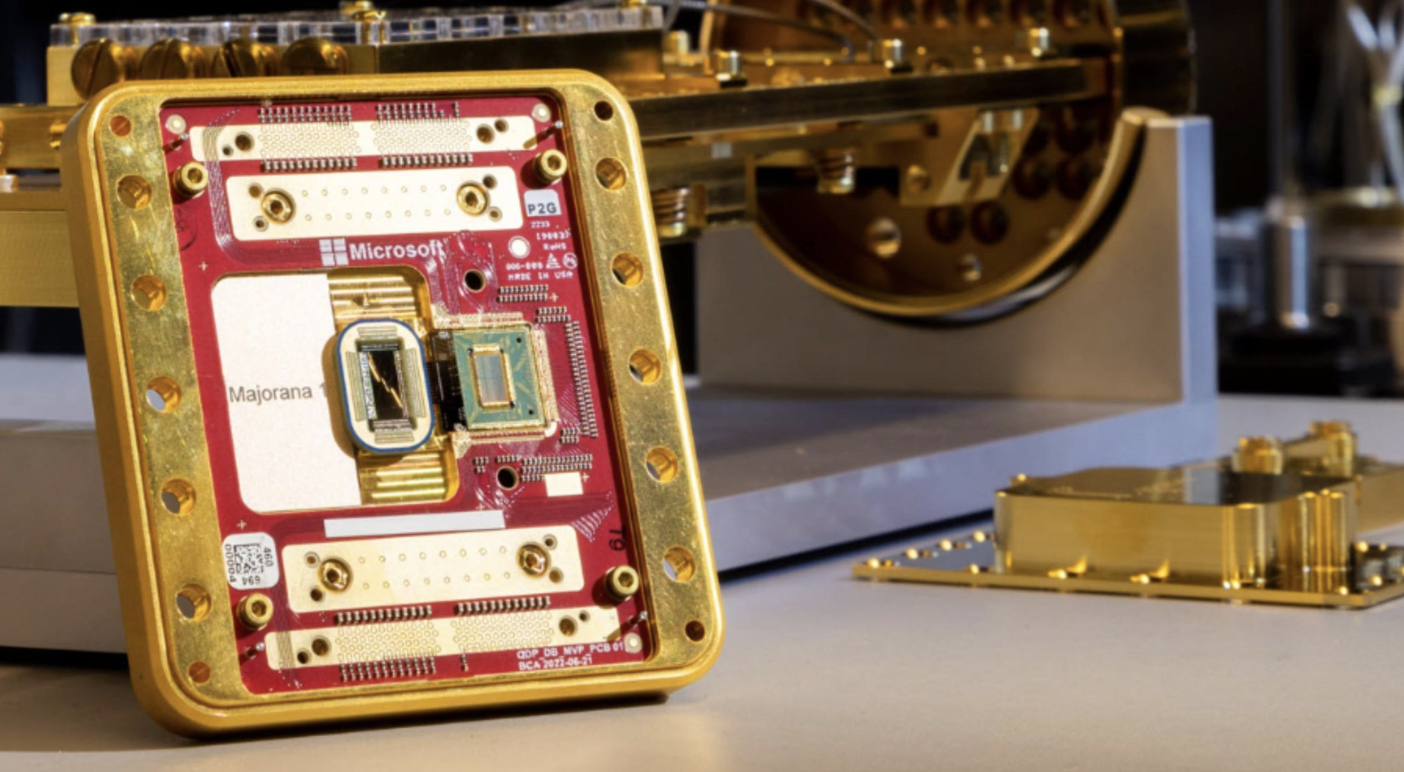 A close-up shot of a small, square, gold-plated, open casing revealing a red circuit board that is marked with the words "Microsoft" and "Majorana". The board holds two small, distinct chips in the center. In the background, out of focus, is a large, complex golden scientific instrument, possibly a cryostat or a quantum computing component.