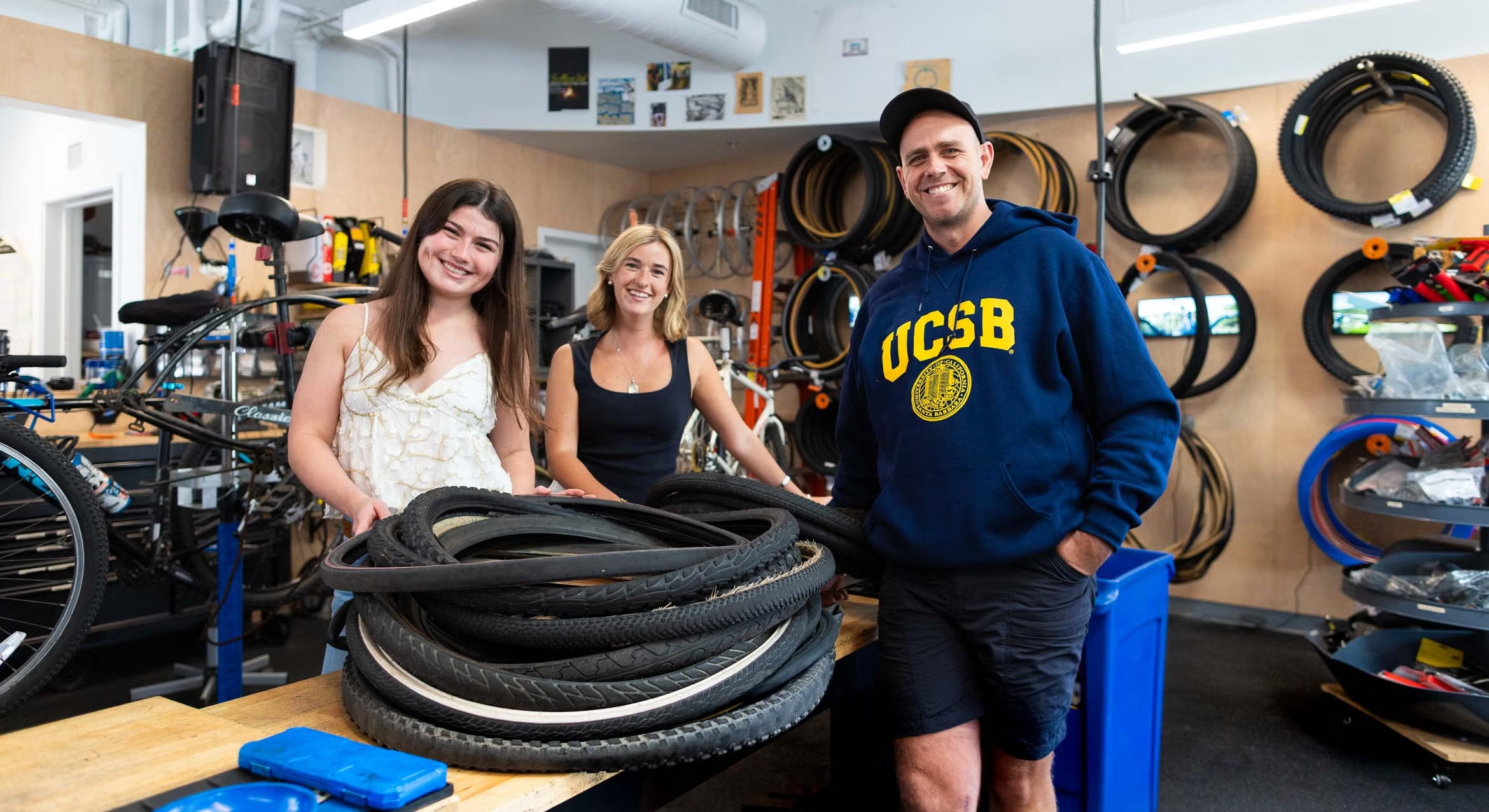 Two female UC Santa Barbara students and the manager of the campus bike shop smile while standing behind a large pile of old bicycle tires.