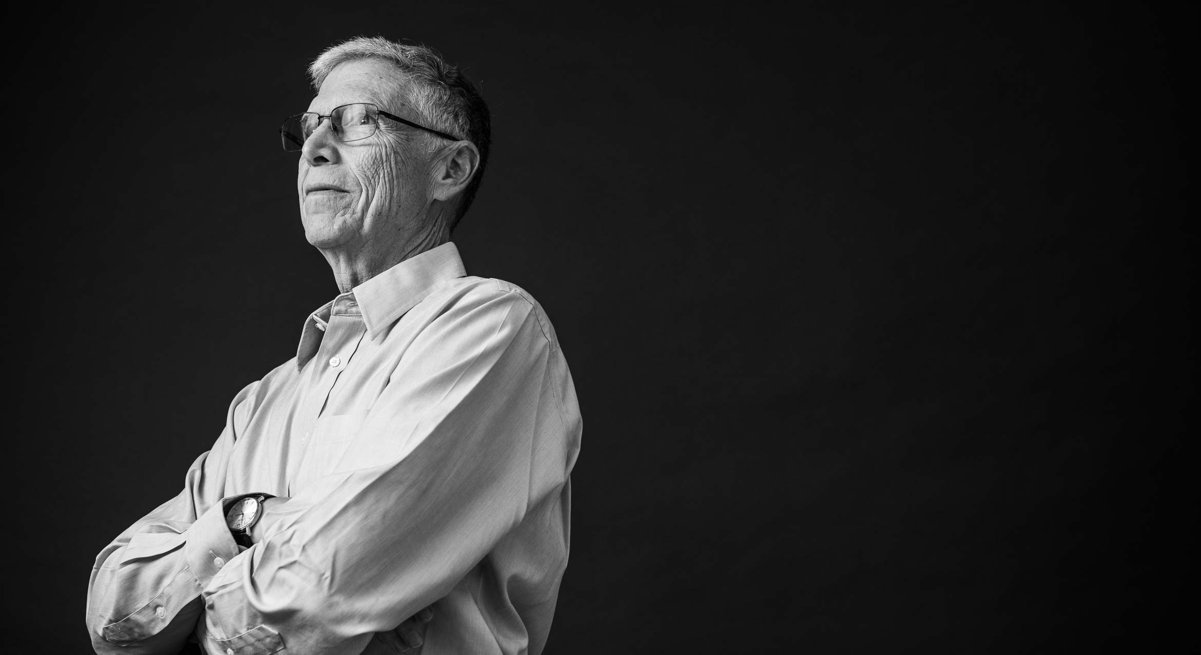 Black and white image of Gary Horowitz with short hair, wearing glasses and a long-sleeved button-up shirt, standing with arms crossed in front of a dark, featureless background.