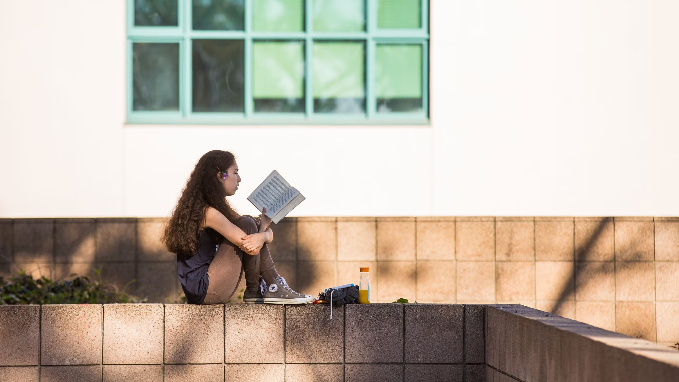 Side profile of a female student with long dark hair sitting cross-legged on a low brick wall, reading a book outdoors with a white building in the background.