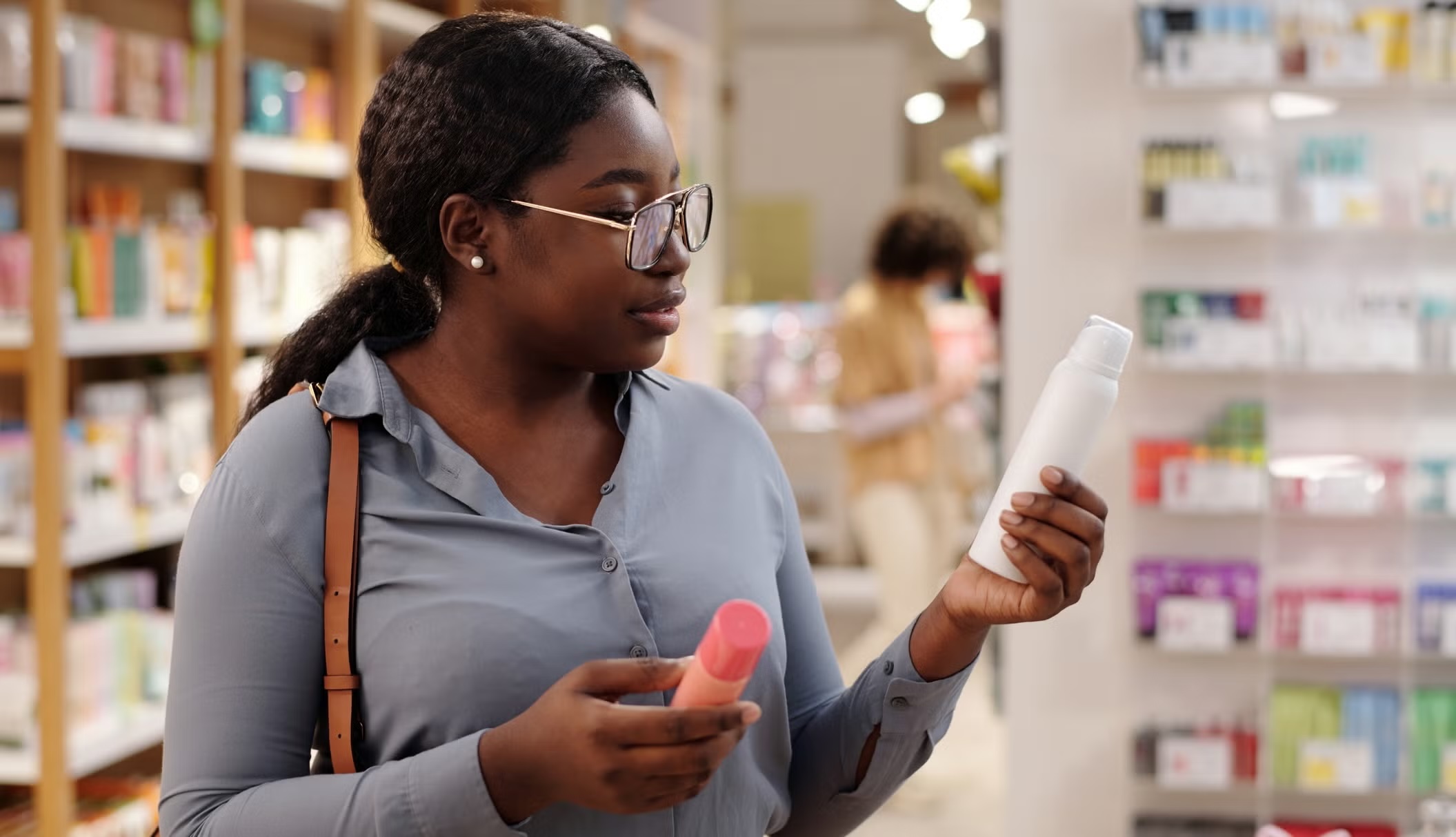 A young Black woman with glasses and her hair in a low ponytail stands in a store aisle, carefully examining a white aerosol bottle in her left hand while holding a small pink and orange container in her right hand.