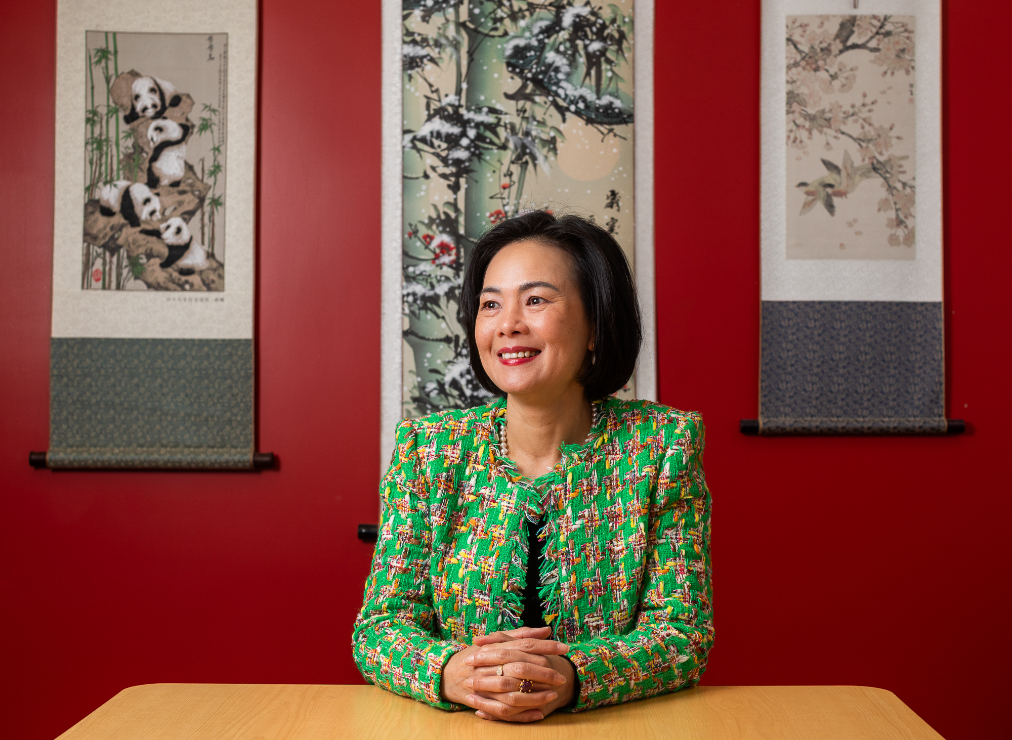 Portrait of Thuc-Quyen Nguyen sitting at a desk. She wears a green tweed jacket against a red wall displaying three traditional scroll paintings.