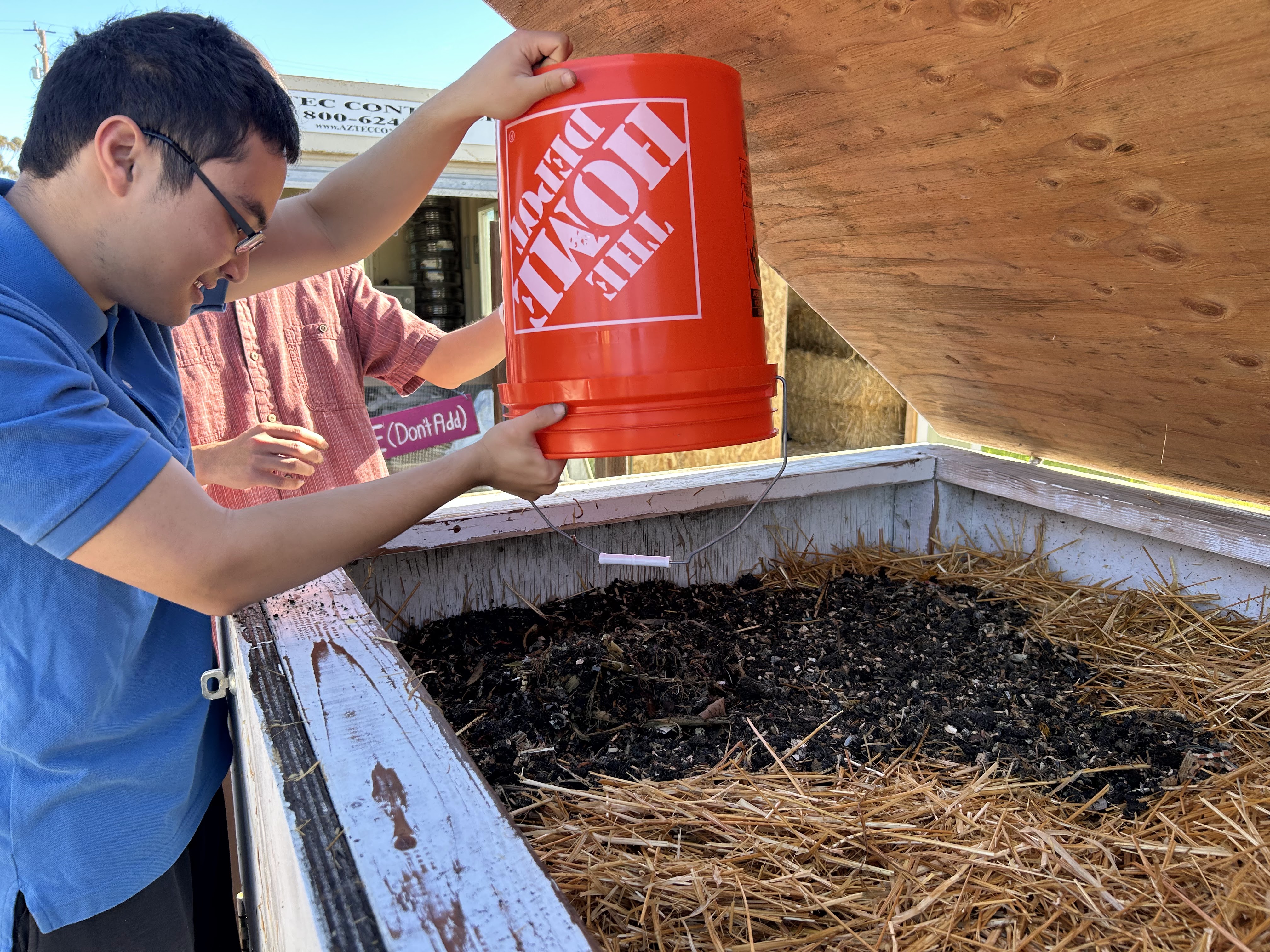 Ben Wollack pouring compost from an orange Home Depot bucket into a wooden compost bin outdoors.