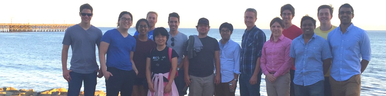 A group of fourteen university students and faculty stand together on a dirt path next to the ocean. The group is diverse in gender and ethnicity. Behind them, the blue water meets a rocky embankment, and a long wooden pier stretches into the distance on the left. The photo is taken during the golden hour, suggesting a team outing or field trip.