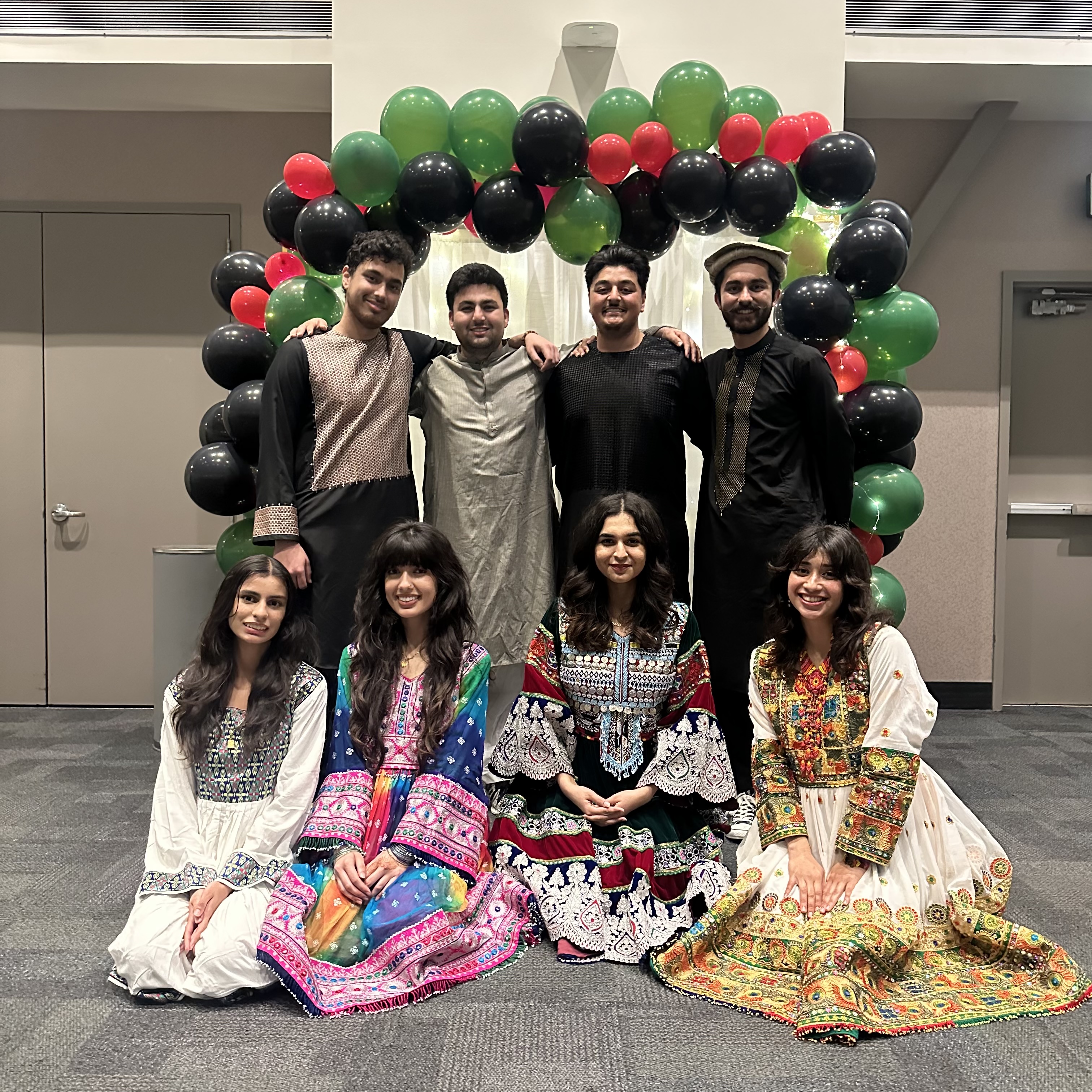 Seven members of the Afghan Student Association posing together in vibrant, traditional Afghan clothing. They are sitting and standing in front of a balloon arch made of black, red, and green balloons.