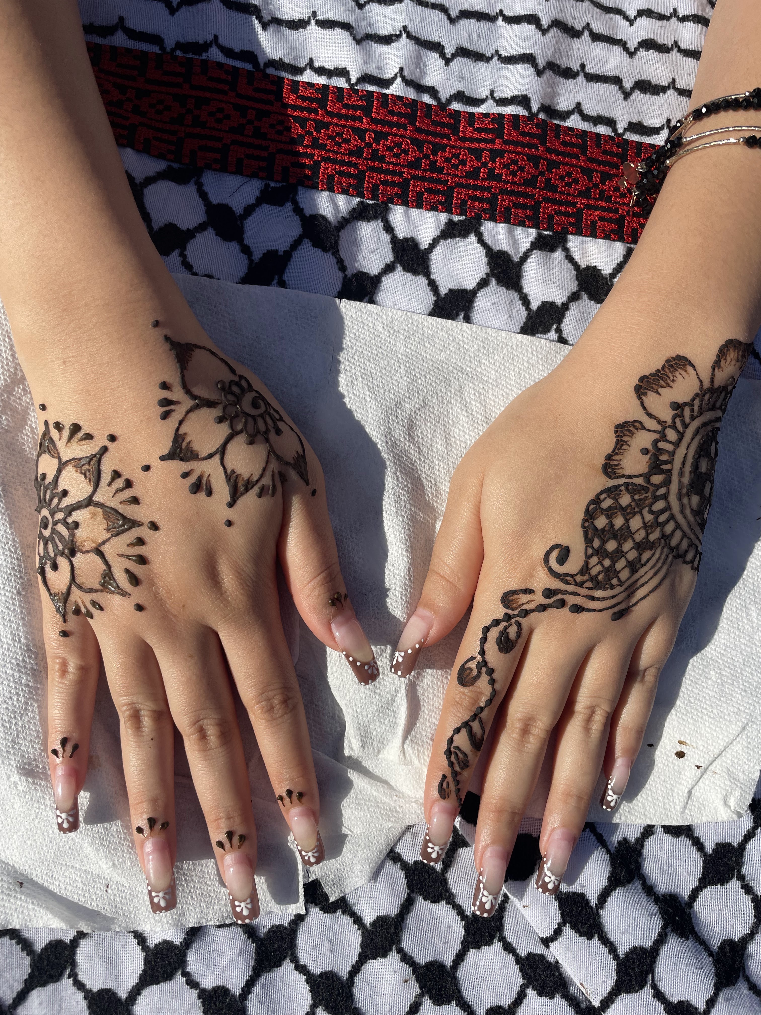 Two hands resting on a white cloth over a black, white, and red patterned textile. The hands feature intricate, dark brown henna floral designs on the backs and fingers, complementing french-tipped nails with small white flowers.