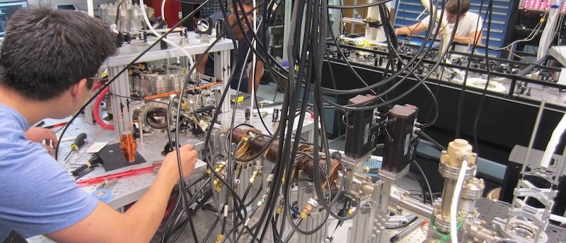 The image shows two individuals, likely students or researchers, working in a cluttered, high-tech laboratory setting. The main subject is a person in a blue shirt intensely working on a complex scientific apparatus, which consists of many metal components, optical tables, and a dense tangle of black cables and tubing. Another person is visible in the background, also working on similar equipment.