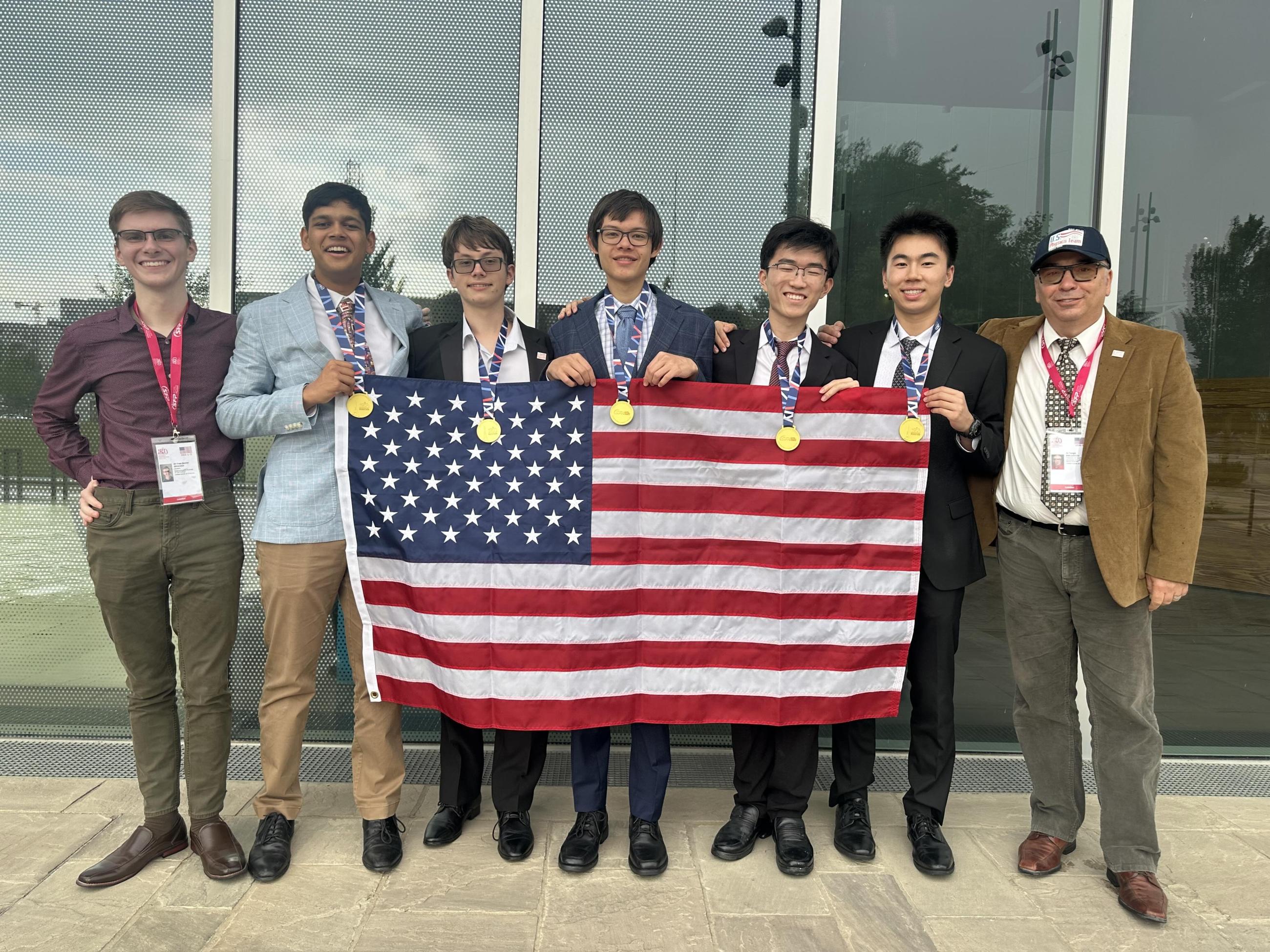 Five students wearing gold medals pose with two coaches and an American flag.