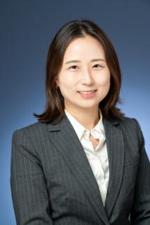Professional headshot of Grace Han against a slate blue background. She is smiling and is wearing a dark gray pinstripe blazer over a white collared shirt.