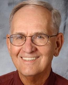 A headshot of Jerry Jacobs smiling warmly. He is wearing wire-rimmed glasses and a maroon button-down shirt.