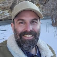 Close-up portrait of Brian Codding with a dark beard and mustache, wearing a tan baseball cap and a green jacket with a white fleece collar, standing in a snowy outdoor setting with a rocky cliff in the background.